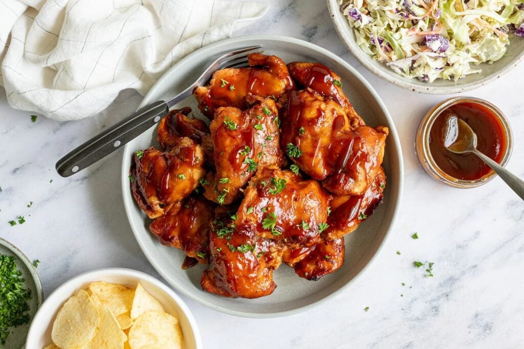 A plate of barbecue chicken thighs garnished with herbs, served with a side of coleslaw, potato chips, and extra barbecue sauce on a marble surface.