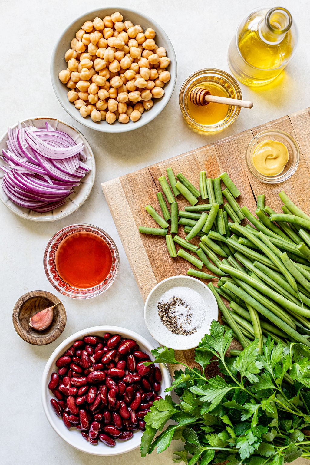 Ingredients for a salad laid out on a counter, including green beans, kidney beans, chickpeas, red onion, parsley, garlic, vinegar, honey, mustard, oil, salt, and pepper.