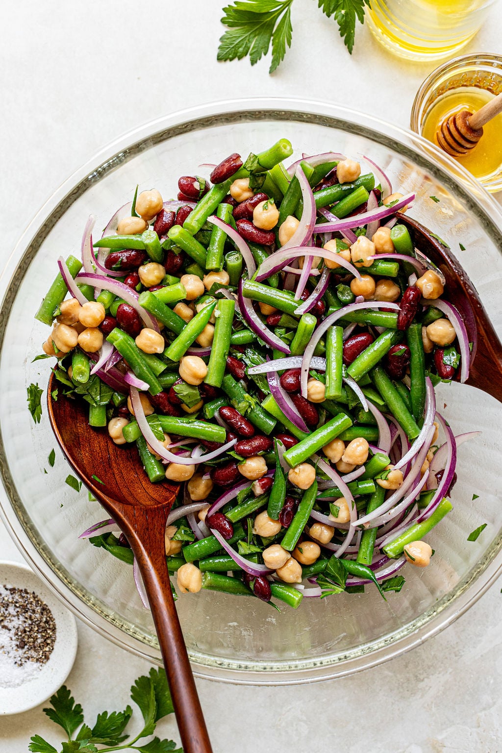 A glass bowl of three bean salad with green beans, kidney beans, chickpeas, sliced red onion, and fresh herbs, with wooden serving spoons, shown on a light surface.