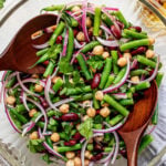 A glass bowl filled with a salad of green beans, kidney beans, chickpeas, red onion, and parsley, with wooden salad servers.