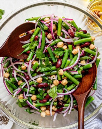 A glass bowl filled with a salad of green beans, kidney beans, chickpeas, red onion, and parsley, with wooden salad servers.