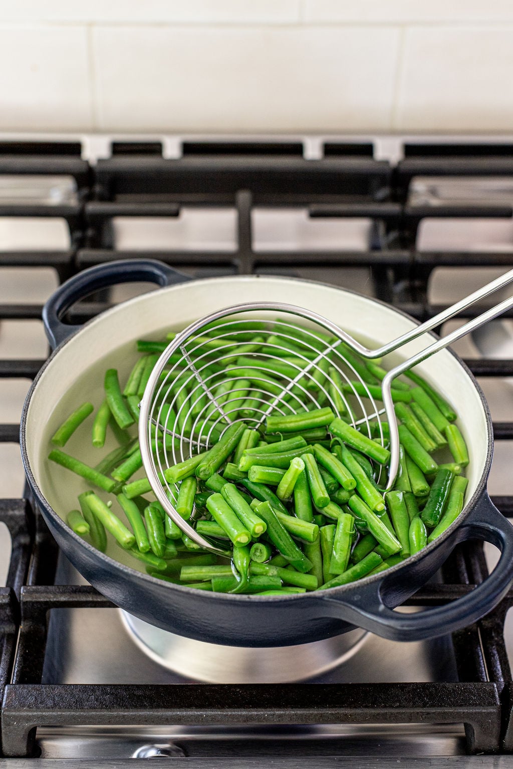 Green beans being blanched in a pot of hot water on a stovetop, with a metal strainer used to lift the beans from the water.