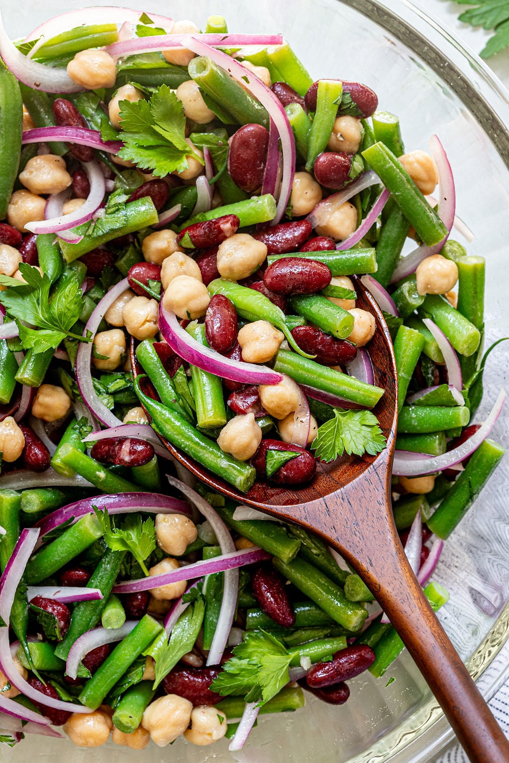 A close-up of a bowl of three-bean salad with kidney beans, chickpeas, green beans, red onion, and parsley, with a wooden spoon serving a portion.