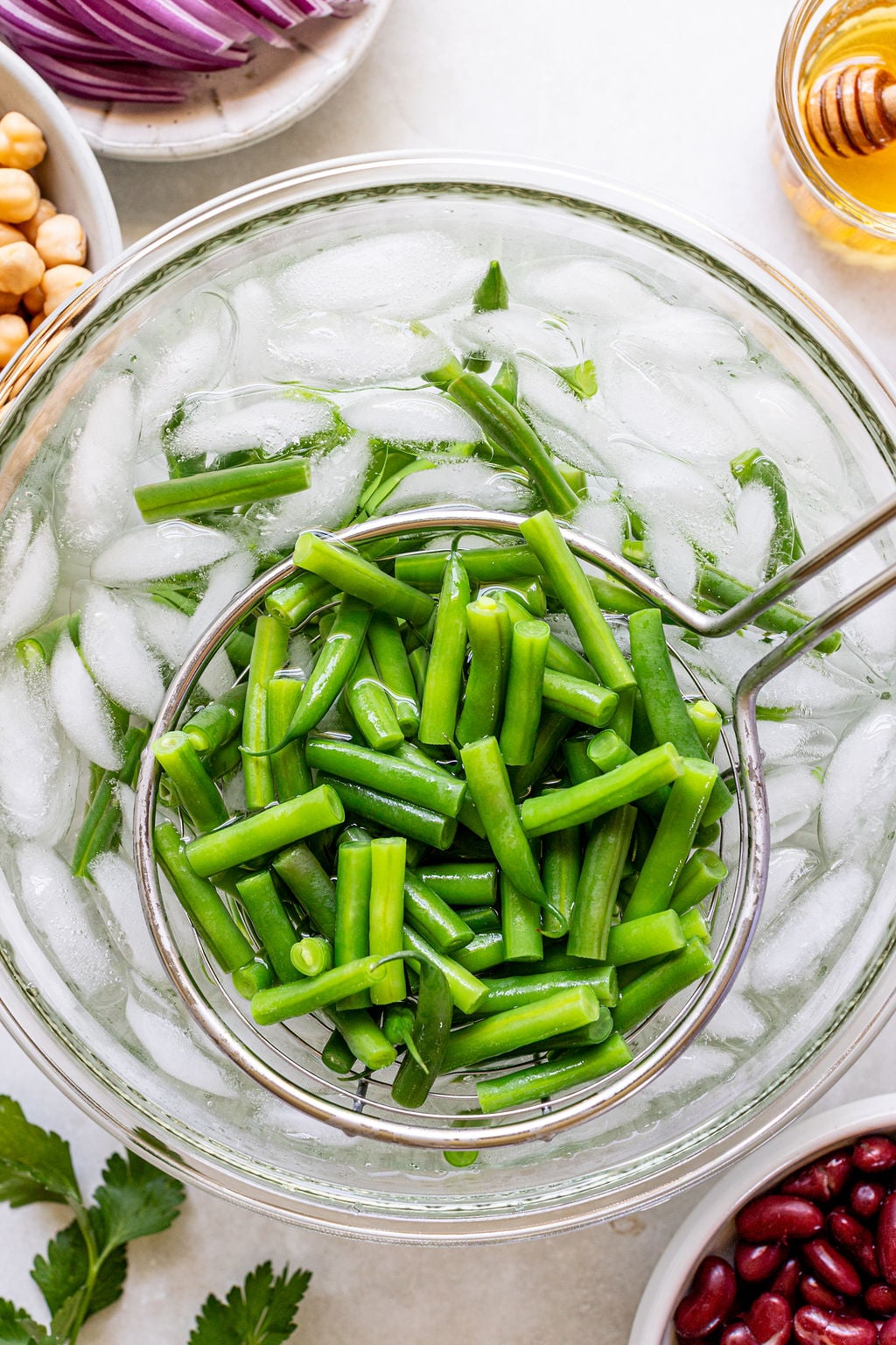 A metal strainer holds blanched green beans over a bowl of ice water, surrounded by ingredients like chickpeas, red onions, parsley, and kidney beans.