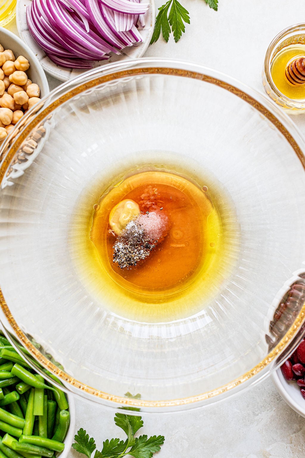 A glass bowl containing olive oil, mustard, salt, pepper, and vinegar sits surrounded by bowls of green beans, chickpeas, red onions, parsley, and honey.