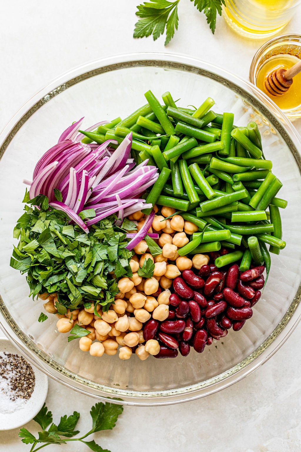 A glass bowl filled with chopped green beans, sliced red onion, chickpeas, kidney beans, and chopped parsley on a light surface.