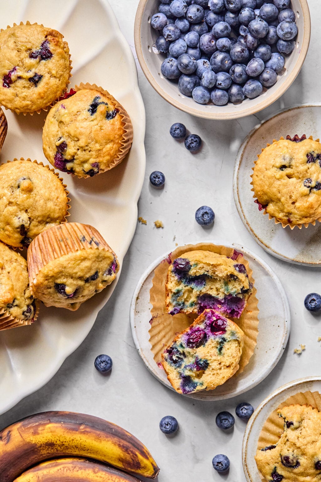 A plate of banana blueberry muffins, a bowl of fresh blueberries, and a pair of ripe bananas on a white surface. One muffin is cut open to show the blueberry-filled interior.