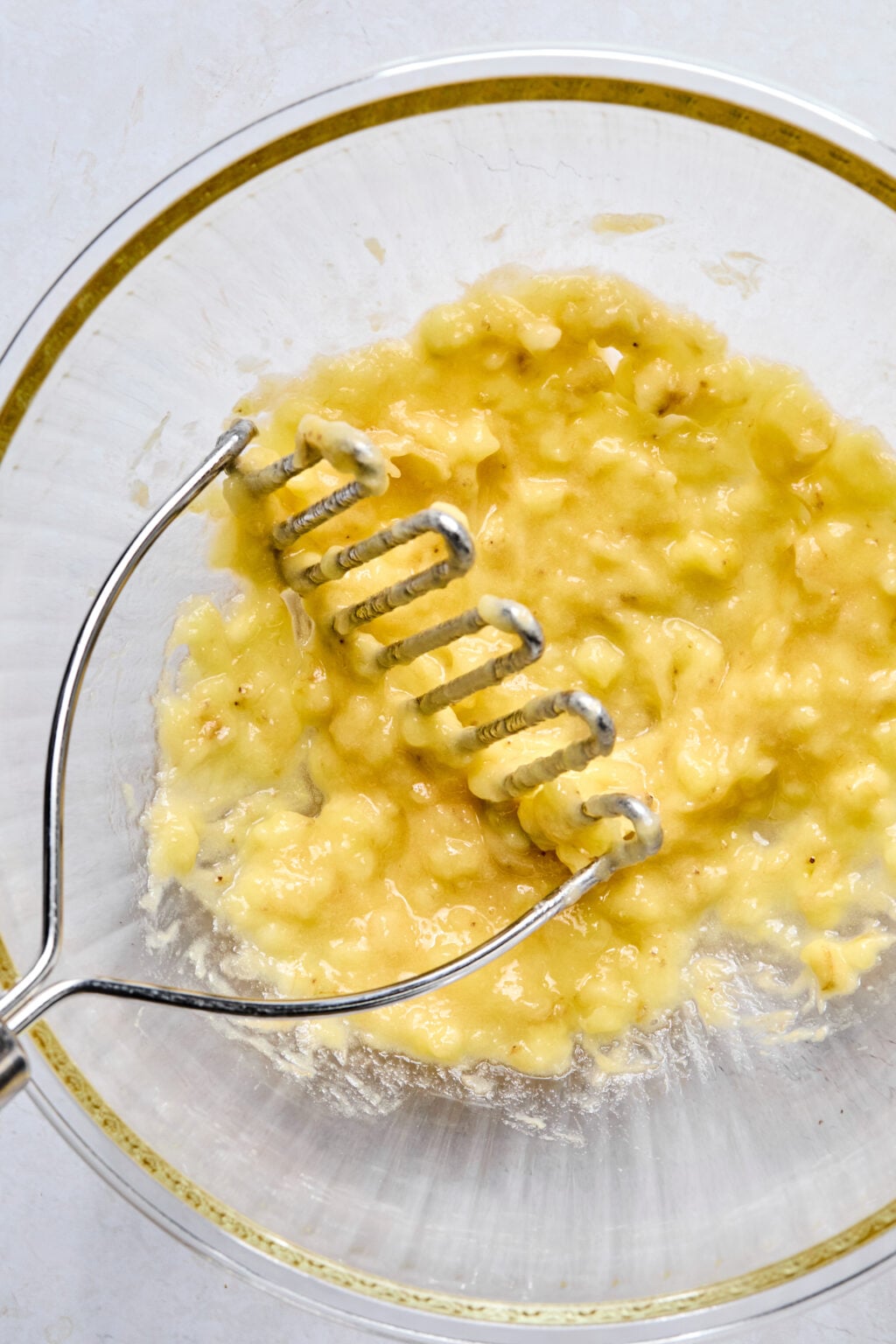 A metal potato masher mashes ripe bananas in a clear glass mixing bowl on a light-colored surface.