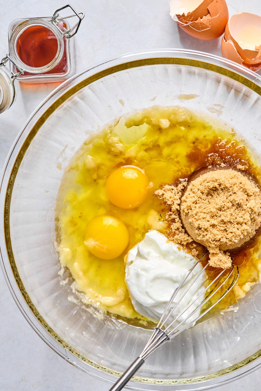 A glass bowl with eggs, brown sugar, yogurt, mashed banana, and vanilla extract ready to be mixed, with a whisk, cracked eggshells, and a jar of vanilla nearby.