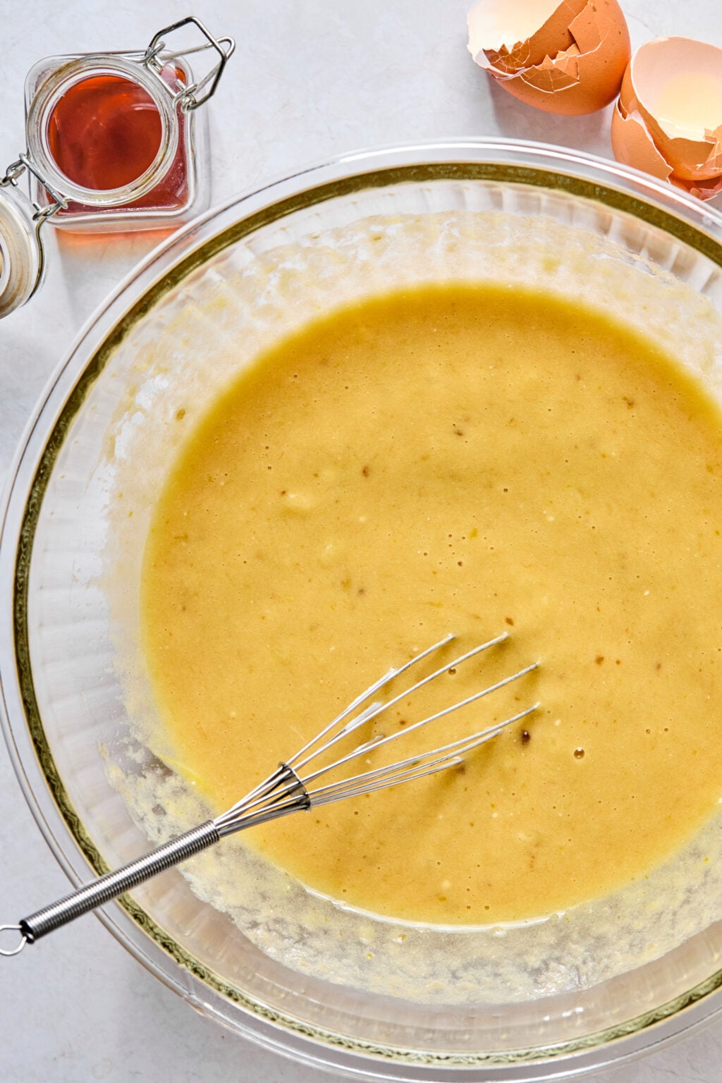 A glass bowl filled with yellow batter and a metal whisk, next to egg shells and a small jar of vanilla extract on a light surface.