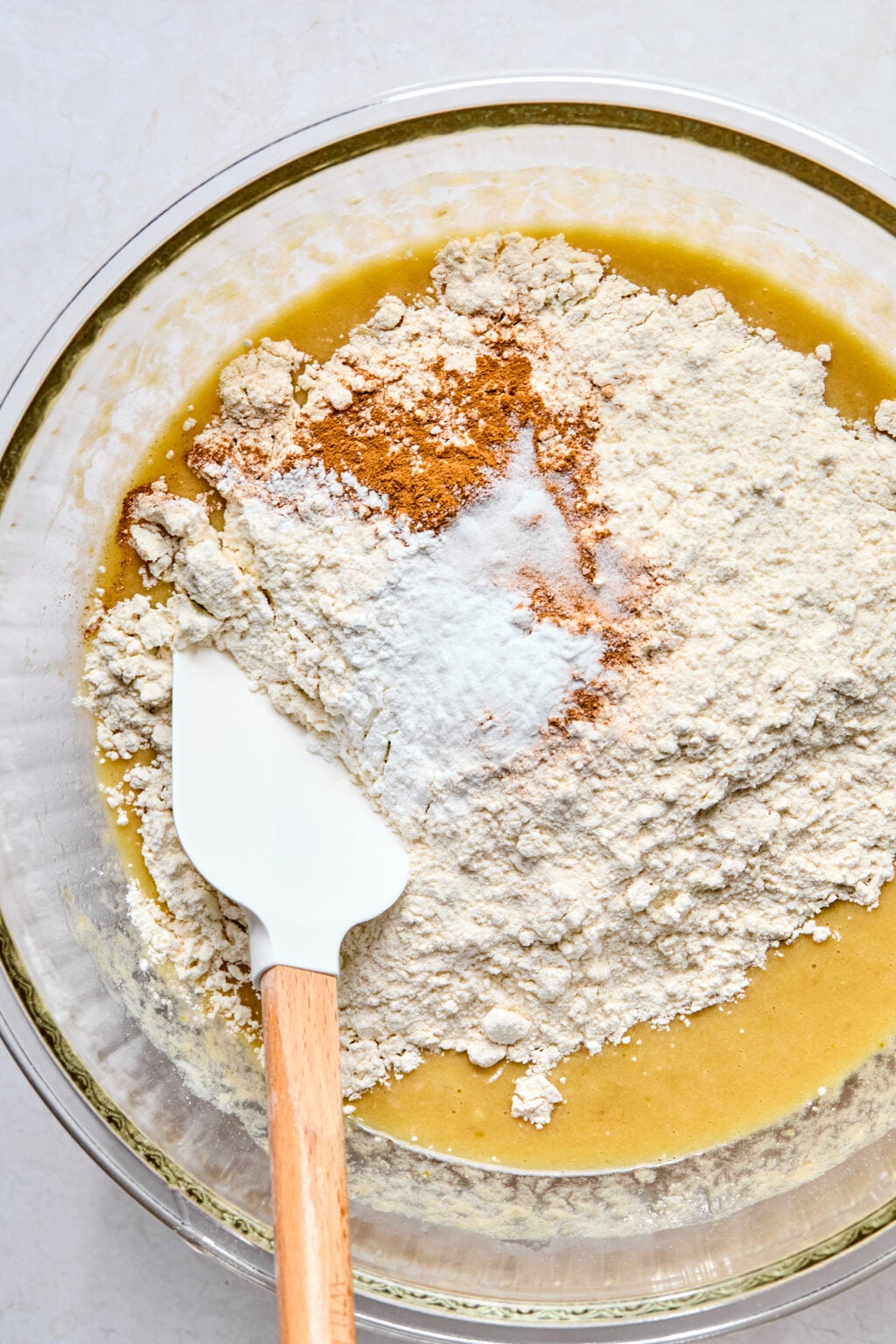 A glass bowl containing flour, baking soda, and spices being mixed into a yellow batter with a white spatula.