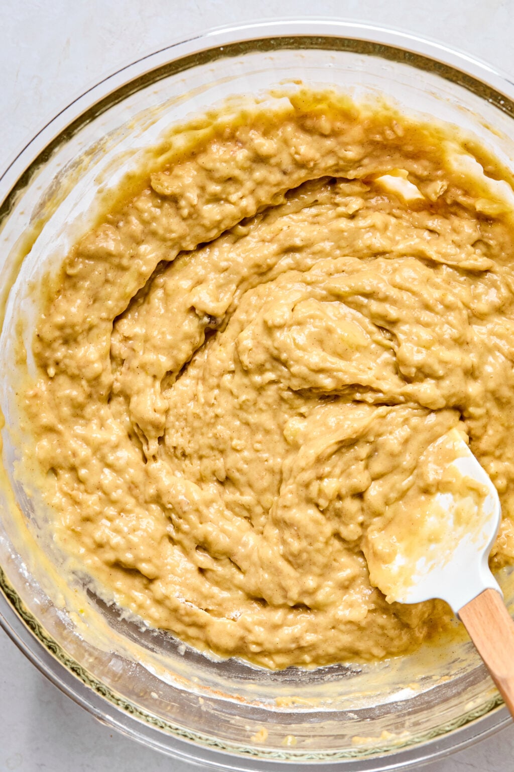 A glass bowl filled with banana muffin batter being mixed with a white spatula.