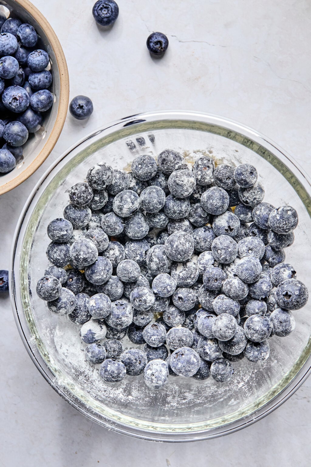 A glass bowl filled with blueberries coated in flour sits on a light surface, with a smaller bowl of plain blueberries nearby.