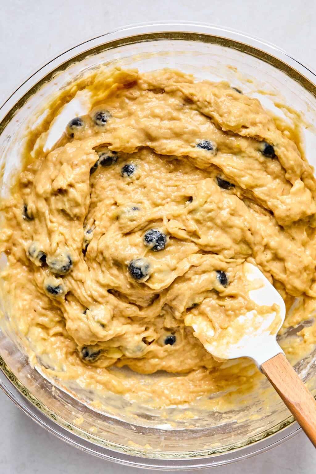 A glass bowl filled with thick, yellowish batter mixed with blueberries, being stirred with a white spatula.