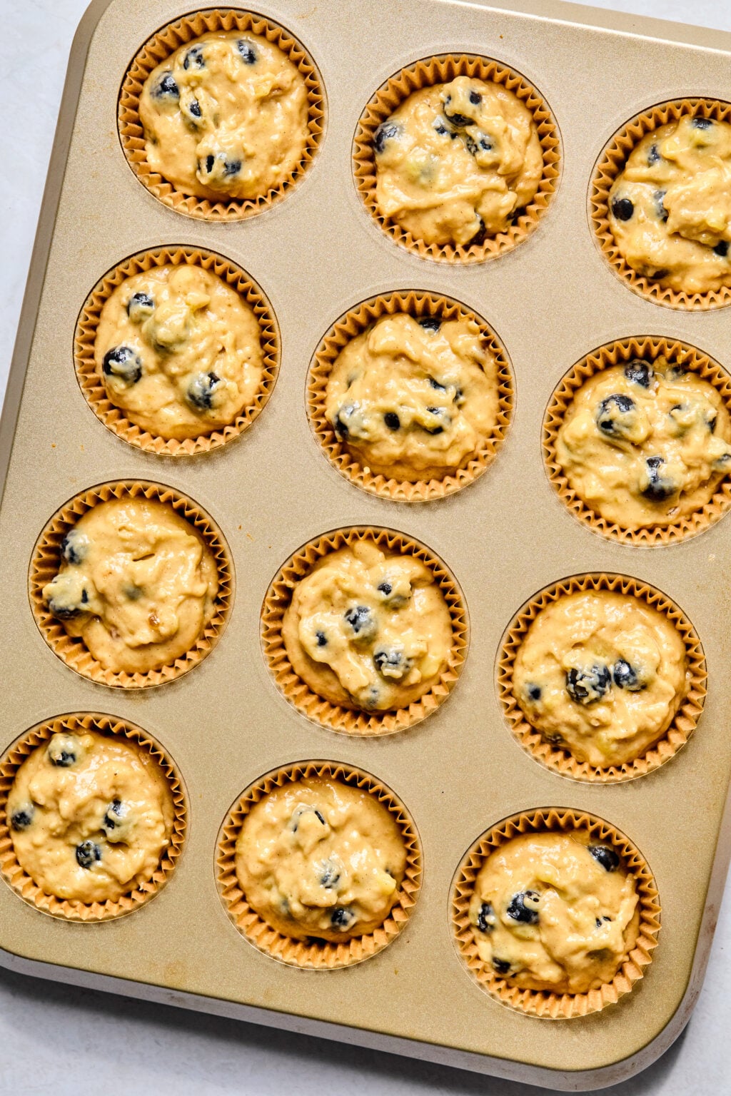A muffin tin filled with twelve paper liners, each holding unbaked muffin batter with visible blueberries.
