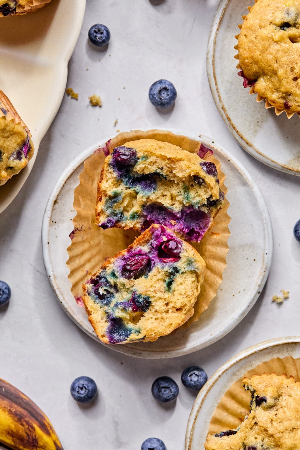 A banana blueberry muffin cut in half on a small plate, surrounded by whole blueberries and other muffins on a light surface.