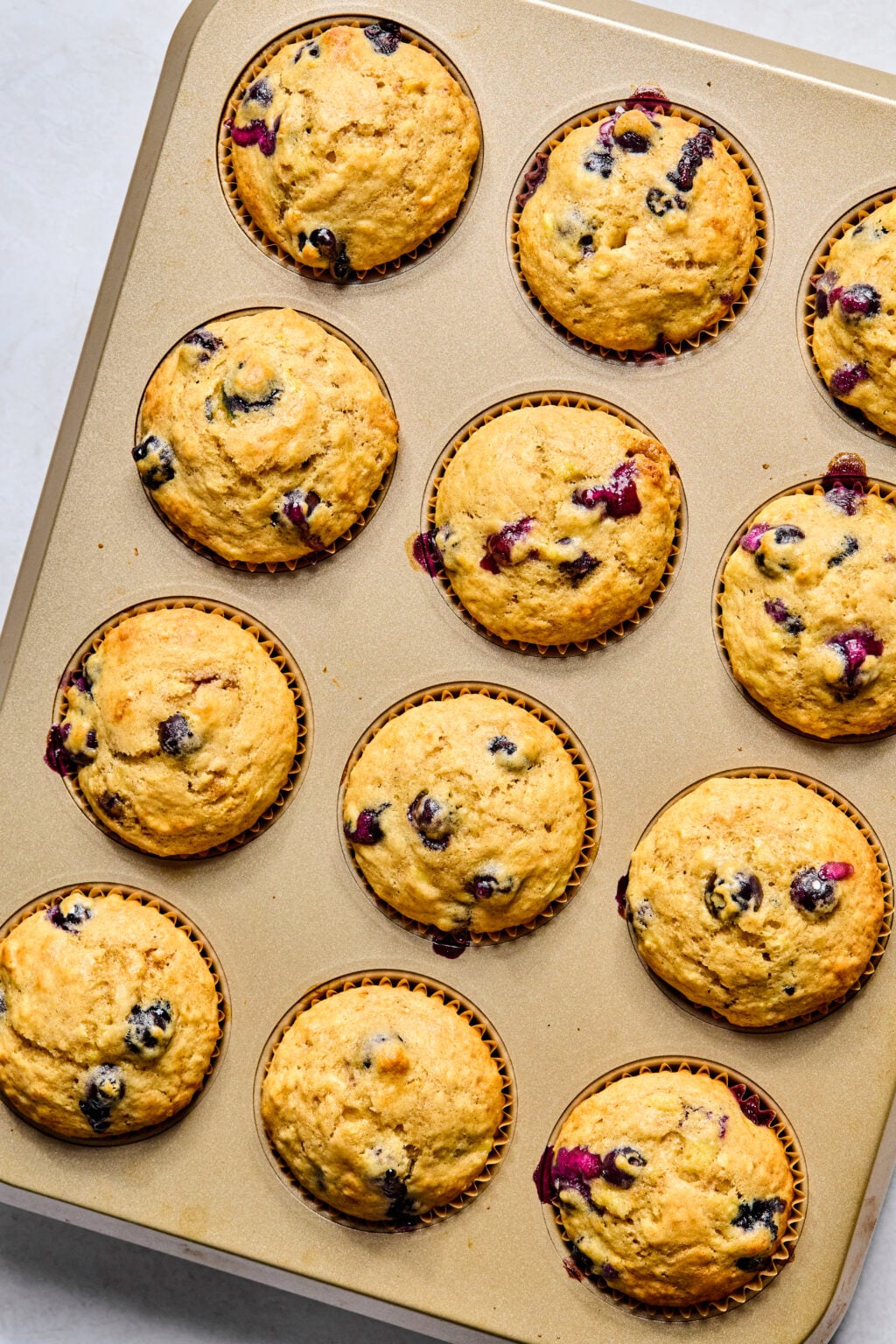 A muffin tin filled with twelve baked blueberry muffins, some with visible blueberries and golden brown tops.