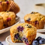A banana blueberry muffin with a bite taken out sits on a plate next to fresh blueberries, with more muffins in the background.