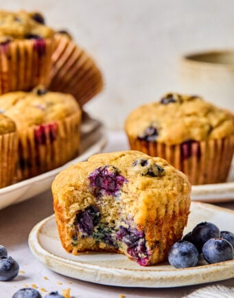 A banana blueberry muffin with a bite taken out sits on a plate next to fresh blueberries, with more muffins in the background.