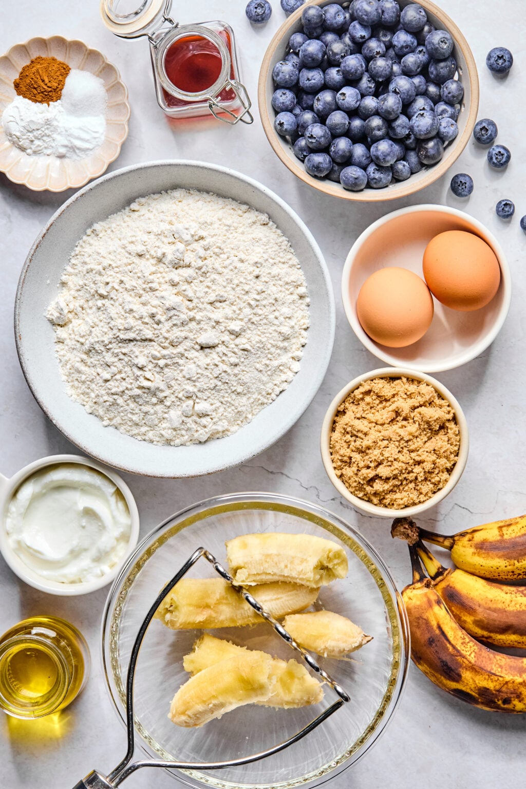Ingredients for baking include flour, brown sugar, eggs, blueberries, bananas, yogurt, oil, vanilla, baking powder, baking soda, and cinnamon arranged on a countertop.