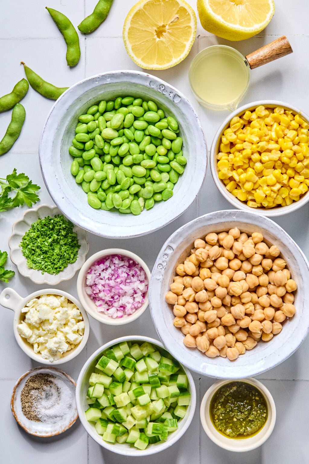 Bowls of edamame, corn, chickpeas, diced cucumber, chopped red onion, feta, parsley, lemon zest, lemon halves, lemon juice, pesto, salt, and pepper arranged on a white surface.
