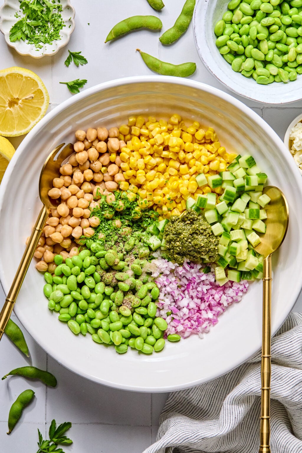 A large white bowl with sections of chickpeas, corn, edamame, diced cucumber, red onion, pesto, and seasonings, with gold serving utensils on the side.
