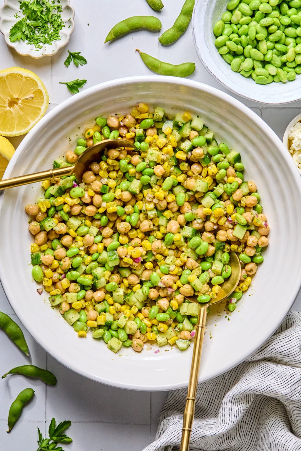 A white bowl filled with a salad made of corn, edamame, chickpeas, and diced cucumber, with two gold serving spoons. Surrounding the bowl are fresh ingredients and a striped cloth.