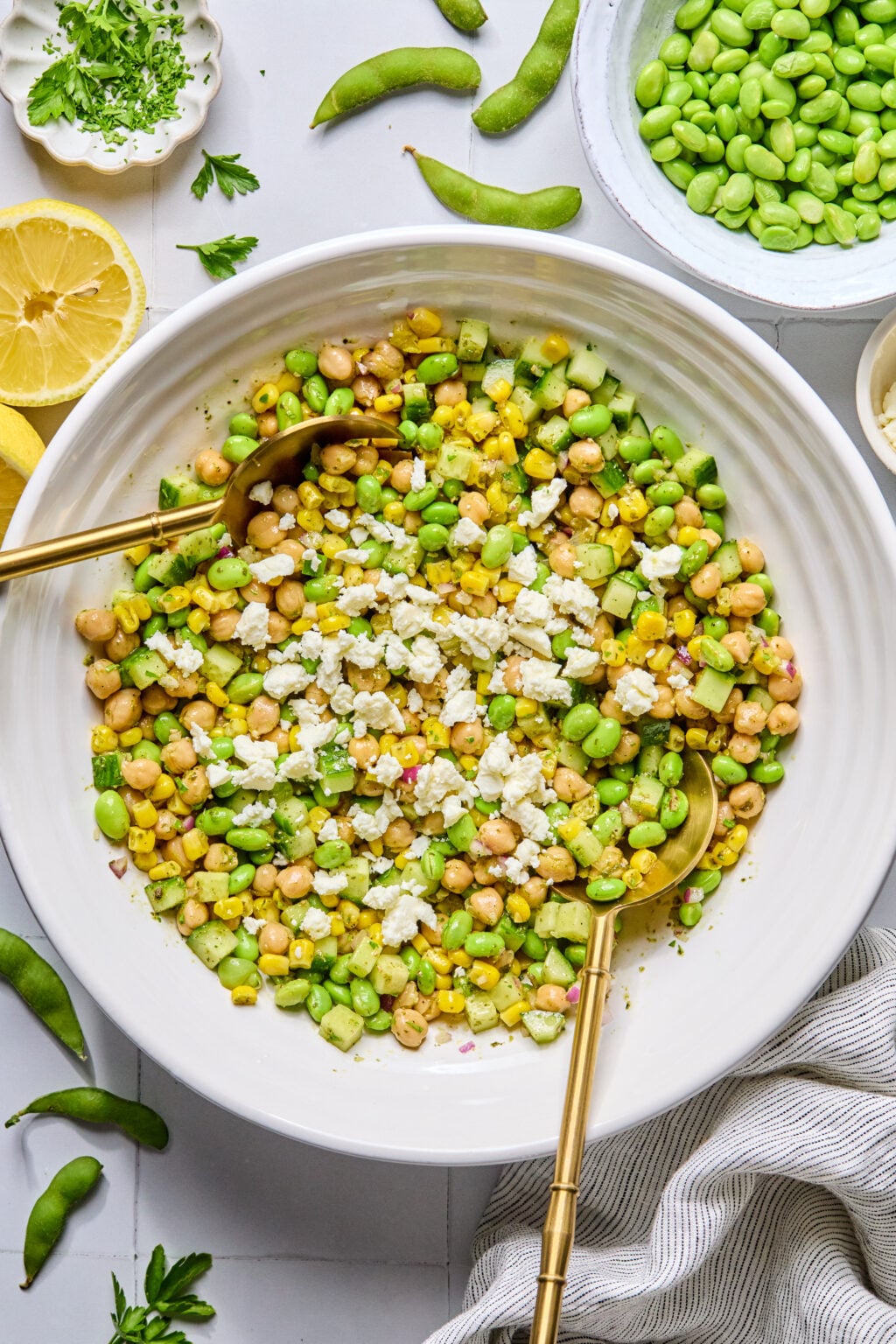 A white bowl contains a salad with corn, edamame, chickpeas, diced cucumber, and crumbled cheese, with two gold serving spoons. Ingredients are scattered around the bowl.