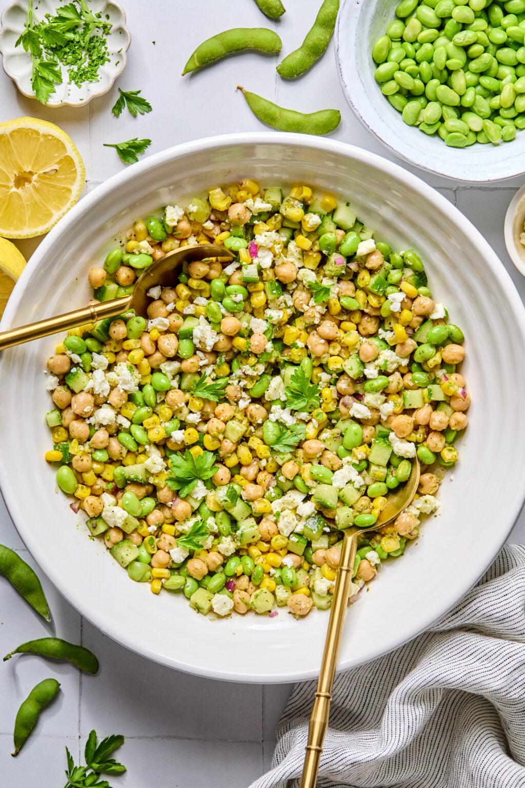 A large white bowl filled with a chickpea, corn, edamame, cucumber, and feta salad, garnished with parsley, with two gold utensils resting inside. Surrounding ingredients are visible.