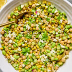 A bowl of chickpea, corn, and edamame salad garnished with fresh parsley and crumbled cheese, served with two golden spoons.