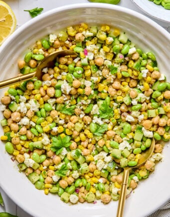 A bowl of chickpea, corn, and edamame salad garnished with fresh parsley and crumbled cheese, served with two golden spoons.