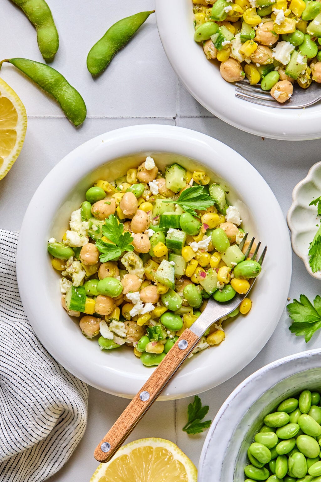 A bowl of salad with chickpeas, edamame, corn, cucumber, feta cheese, and herbs, with a fork resting inside. Lemon slices and edamame pods are nearby on a tiled surface.