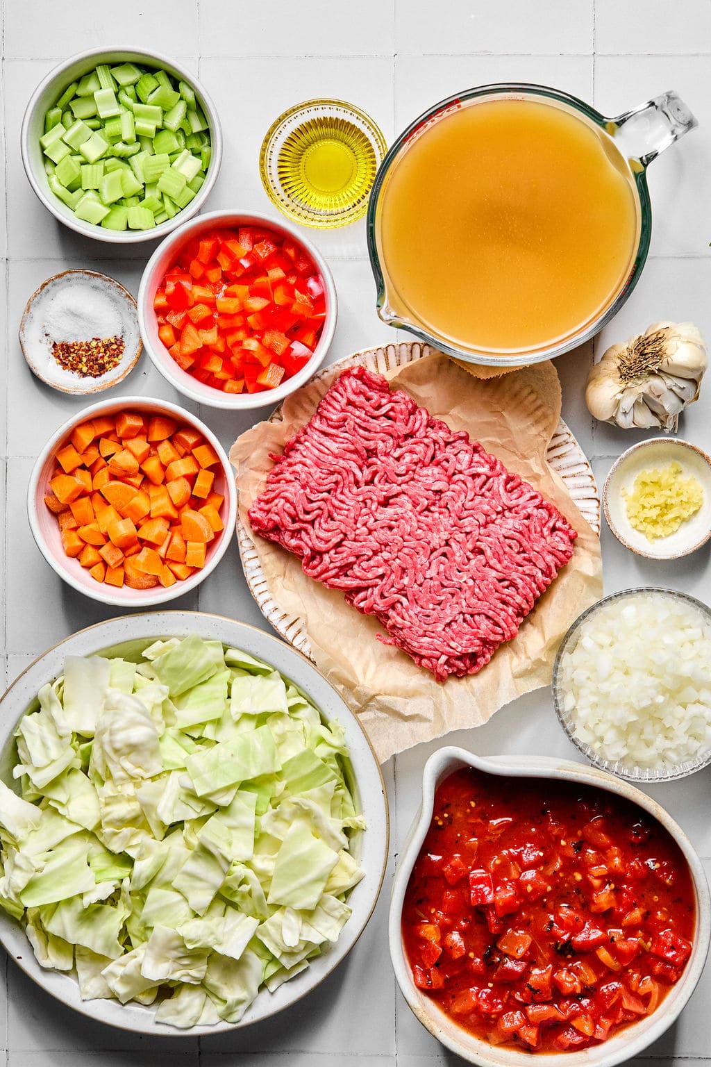 Top-down view of raw ground beef, chopped cabbage, carrots, celery, red bell pepper, onion, garlic, broth, diced tomatoes, olive oil, and seasonings arranged on a white surface.