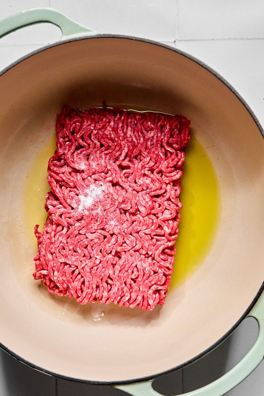Raw ground beef with a sprinkle of salt sits in olive oil in a large light-colored pot on a tiled surface.