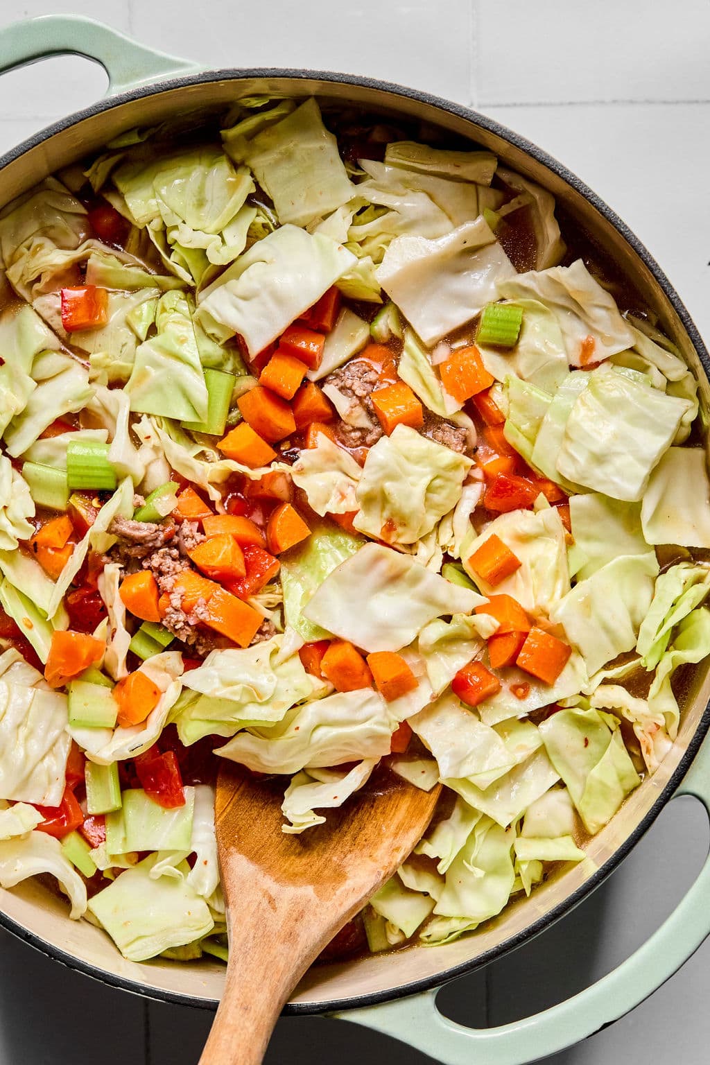 A pot of chopped cabbage, carrots, celery, and tomatoes being stirred with a wooden spoon.