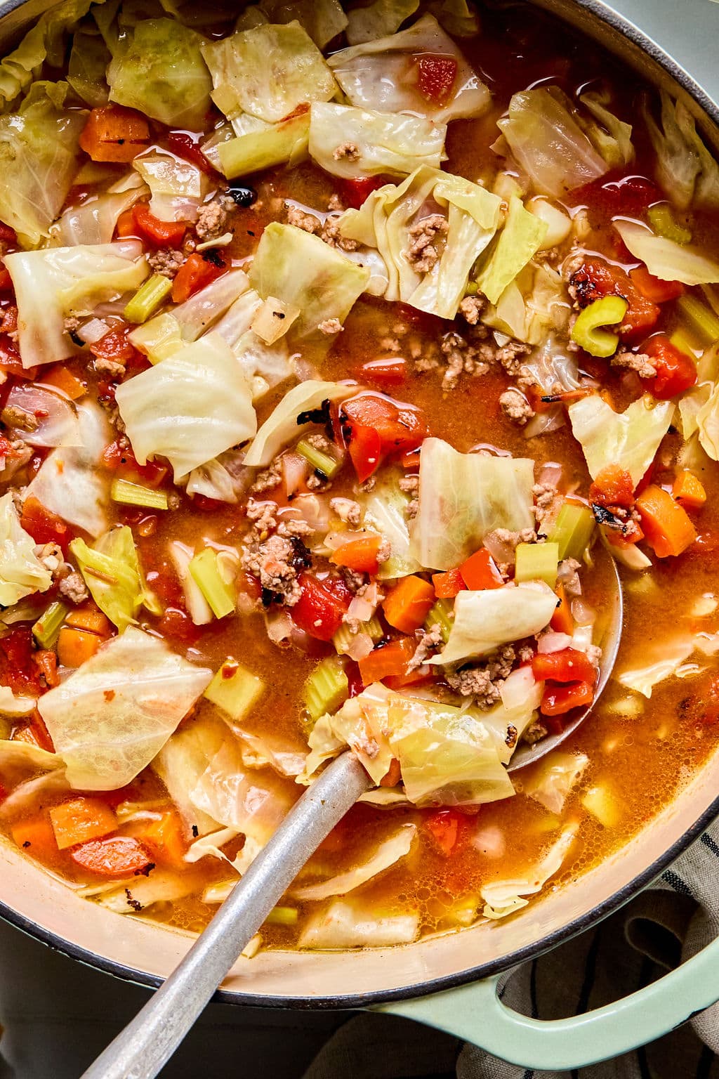 A pot of cabbage soup with ground meat, chopped tomatoes, celery, carrots, and broth, with a ladle submerged in the mixture.