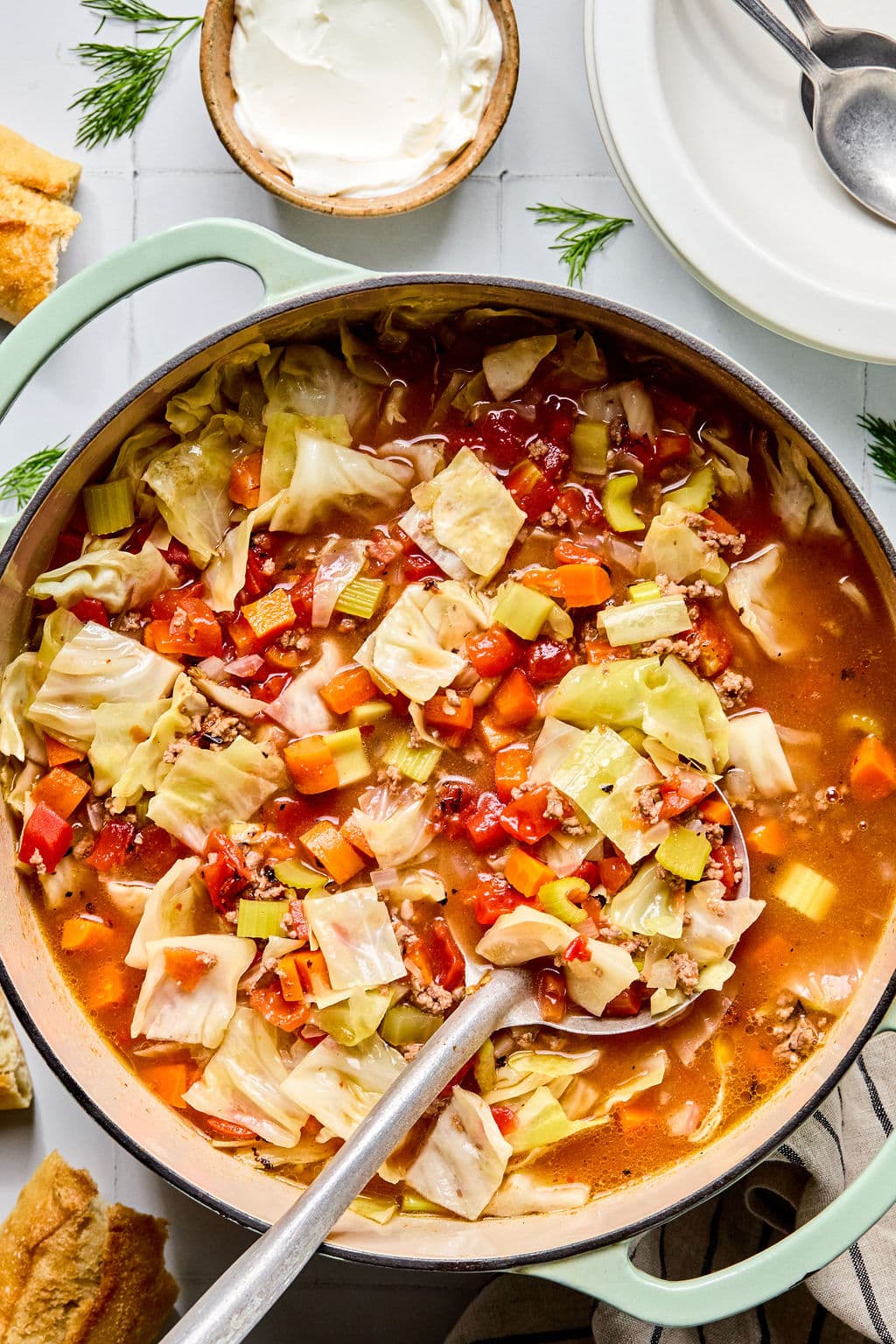 A pot of cabbage soup with chopped cabbage, carrots, celery, tomatoes, and ground meat sits on a stove, with a ladle, bread, and a bowl of sour cream nearby.