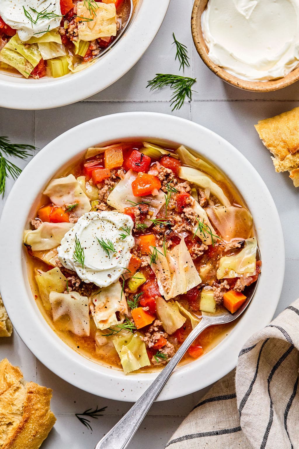 A bowl of cabbage soup with ground meat, carrots, tomatoes, and cabbage, topped with a dollop of sour cream and fresh dill, with bread and extra sour cream on the side.