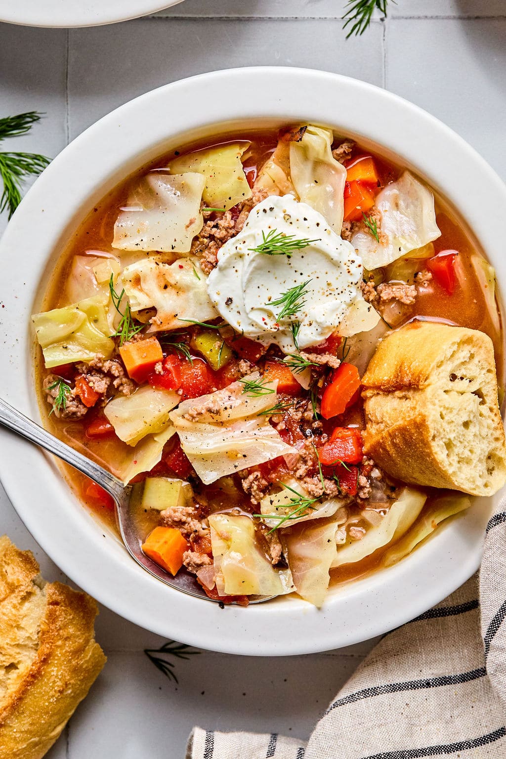 A bowl of cabbage soup with ground beef, carrots, tomatoes, and a dollop of sour cream, garnished with dill, served with a piece of bread.