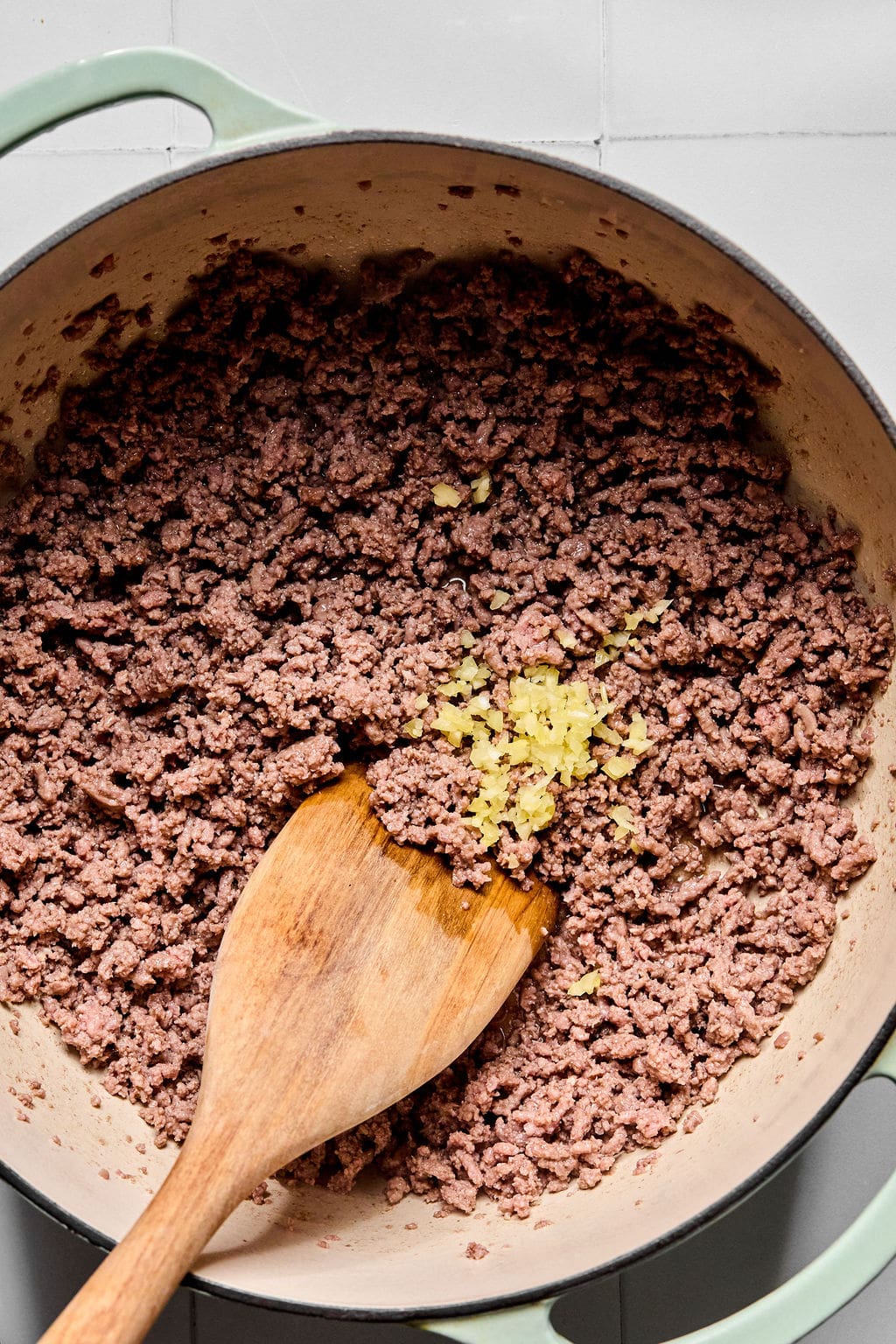 Ground beef is being browned in a pot, with minced garlic added and a wooden spoon stirring the mixture.