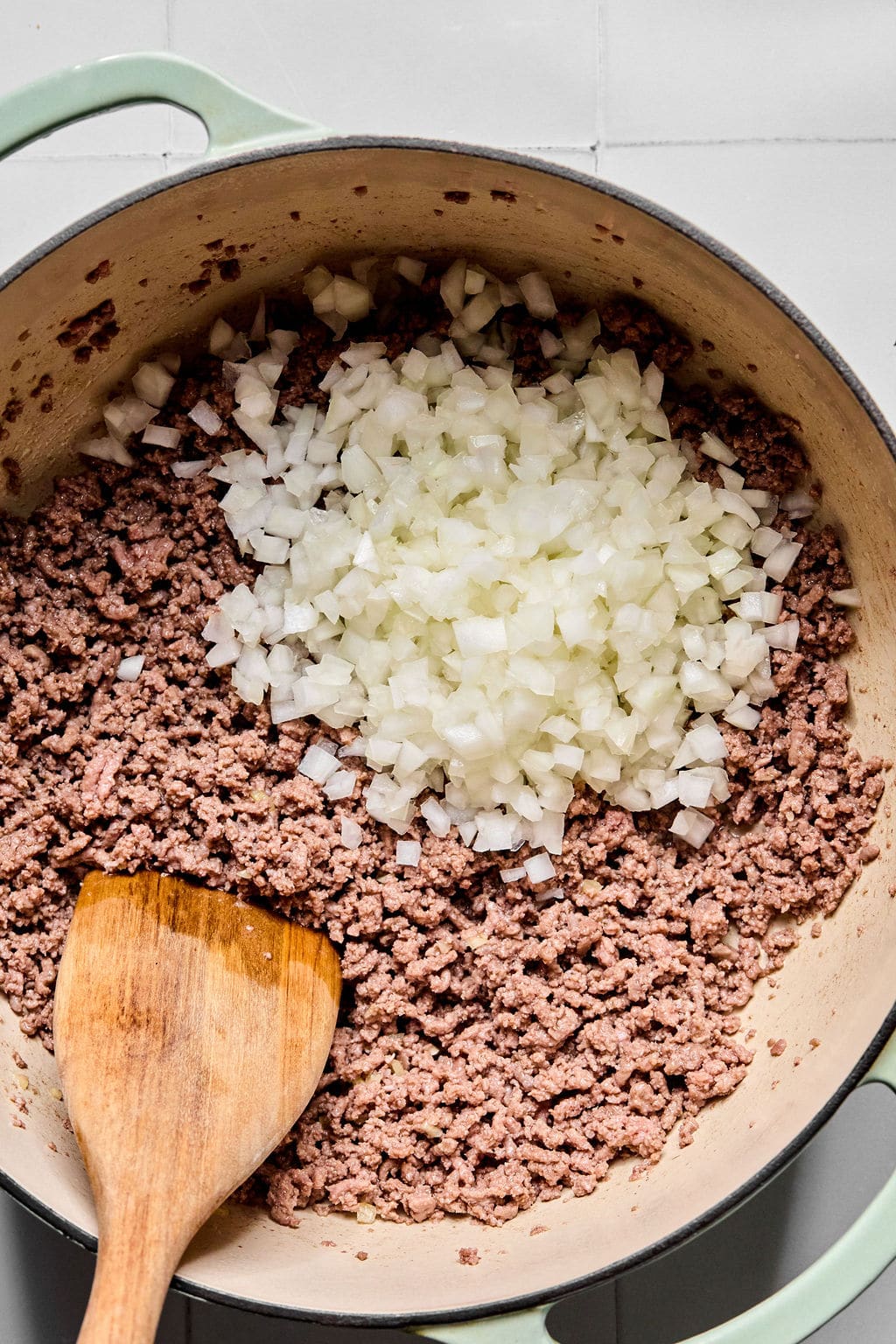 Chopped onions and cooked ground beef in a large pot with a wooden spatula, on a light-colored surface.