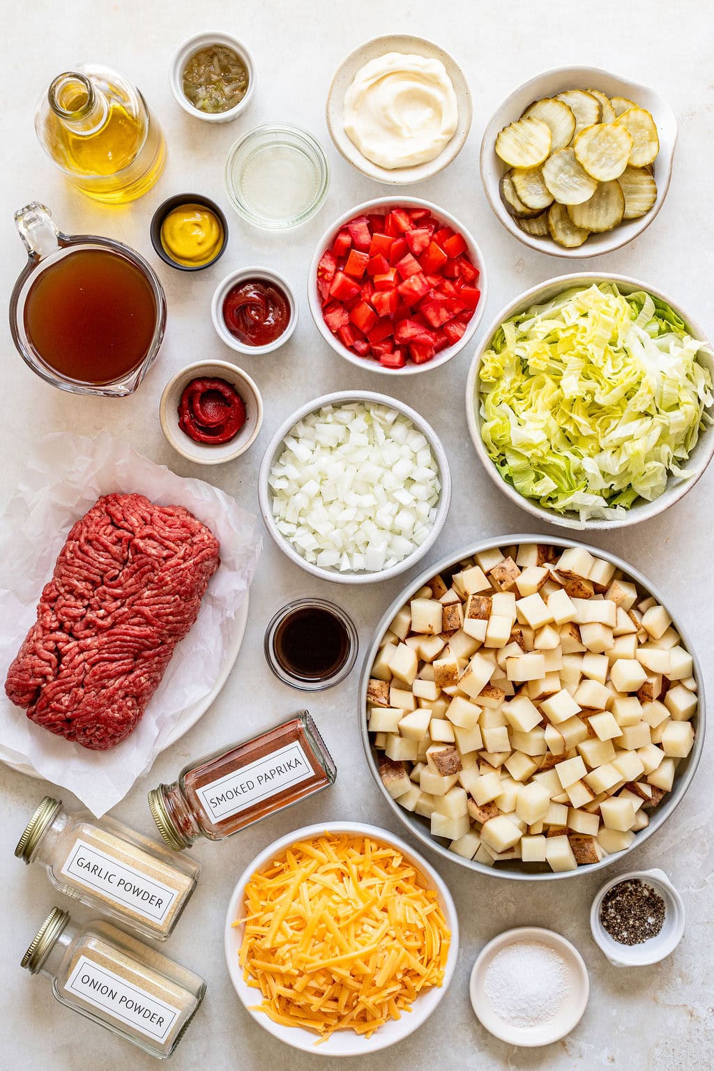 Assorted ingredients on a table, including ground beef, diced potatoes, shredded cheese, lettuce, onions, seasonings, pickles, condiments, chopped tomatoes, broth, and mayonnaise.