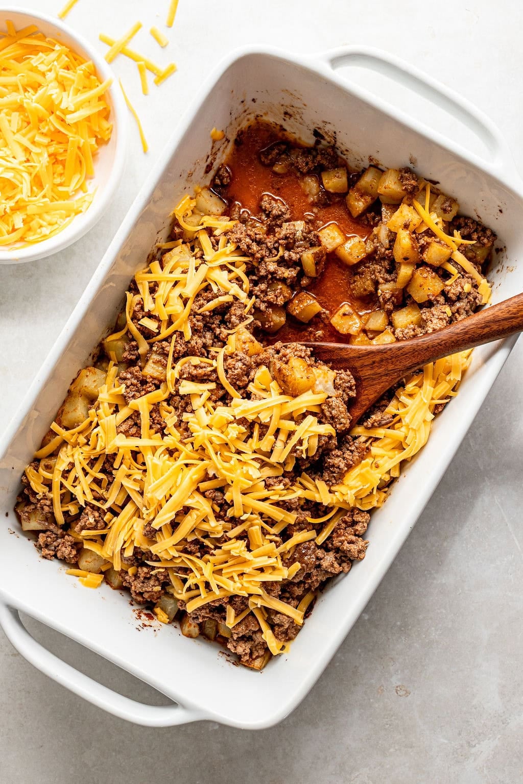 Rectangular baking dish with cooked ground beef, diced potatoes, and shredded cheddar cheese, with a wooden spoon and a bowl of extra shredded cheese on the side.