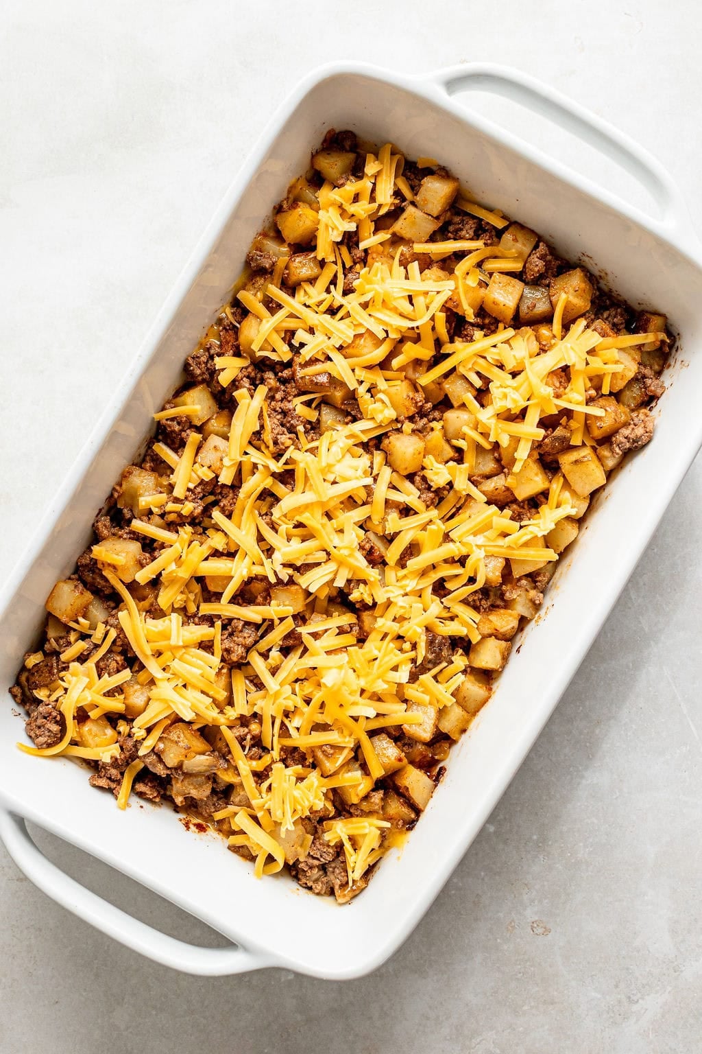 Casserole dish with a layer of ground meat, diced potatoes, and shredded cheddar cheese on top, ready to be baked.