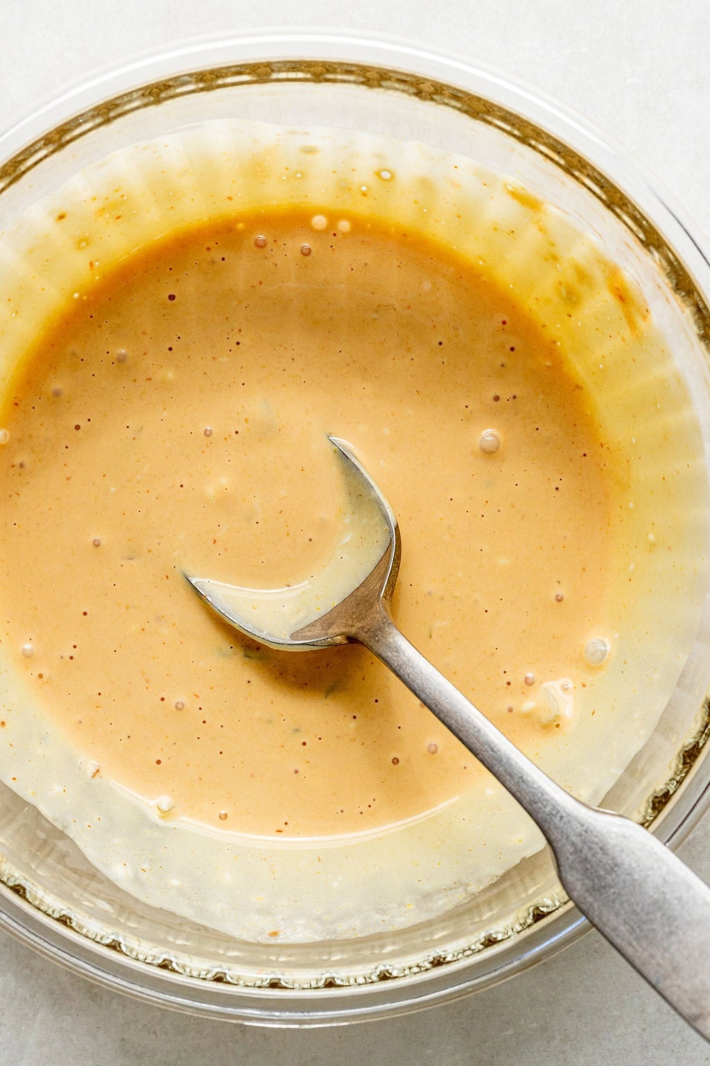 A metal spoon rests in a glass bowl filled with a light brown, creamy sauce.