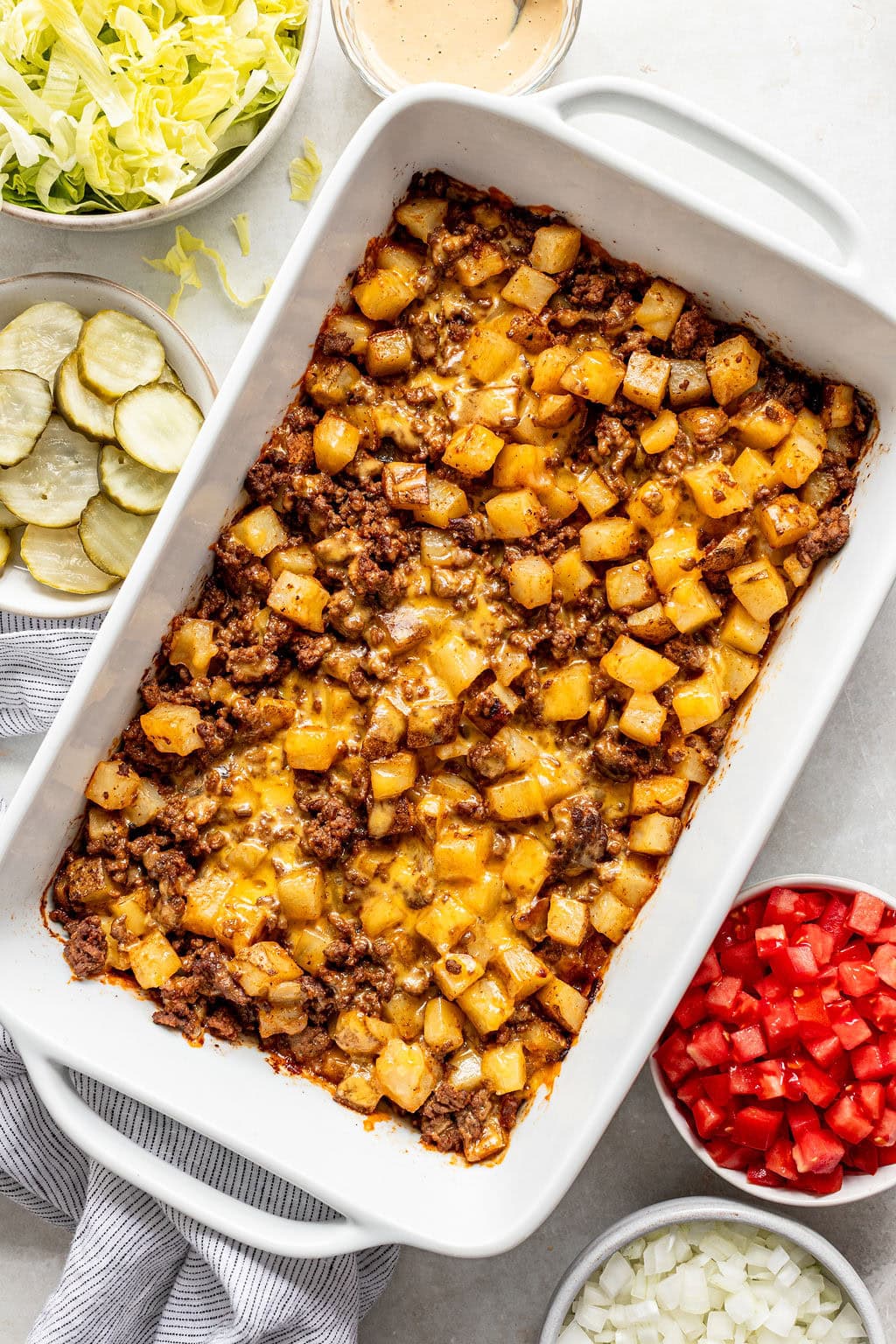 Rectangular casserole dish with baked ground beef, diced potatoes, and melted cheese, surrounded by bowls of lettuce, pickles, tomatoes, onions, and a small bowl of sauce.