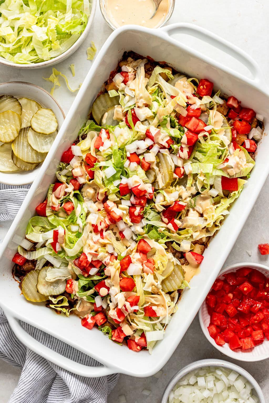 Rectangular dish with a layered casserole topped with shredded lettuce, chopped tomatoes, diced onions, pickles, and a creamy dressing. Bowls of lettuce, pickles, tomatoes, and onions are nearby.