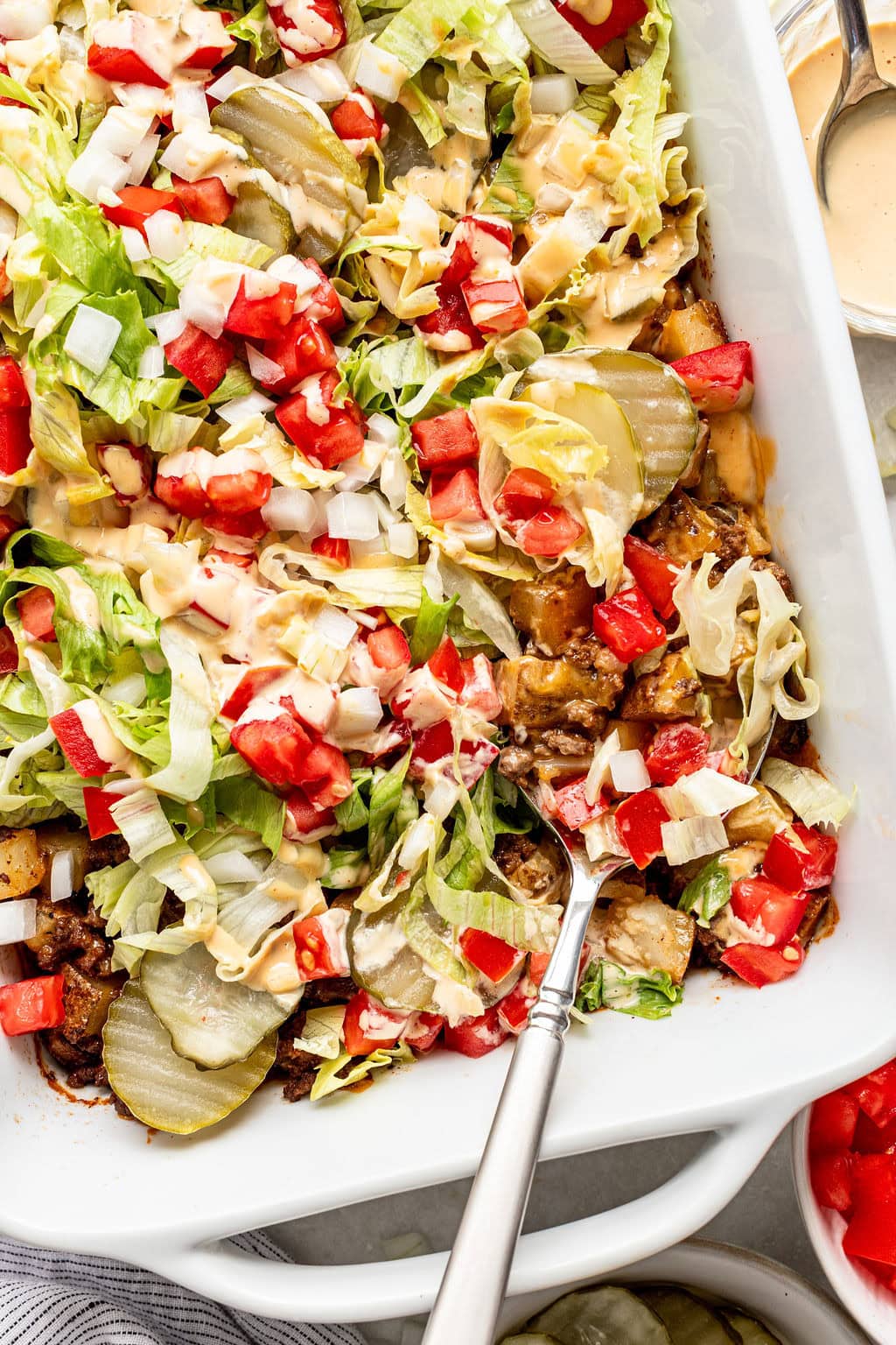 A casserole dish filled with layers of ground beef, lettuce, diced tomatoes, onions, pickles, shredded cheese, and sauce, with a serving spoon inside.