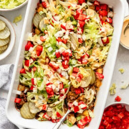 Rectangular white baking dish filled with a layered casserole topped with shredded lettuce, diced tomatoes, onions, cheese, and pickles, surrounded by bowls of the same ingredients.