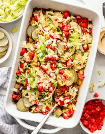 Rectangular white baking dish filled with a layered casserole topped with shredded lettuce, diced tomatoes, onions, cheese, and pickles, surrounded by bowls of the same ingredients.